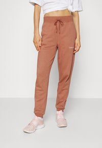 Harmaan ruskeat collegehousut, joissa on nauhavyötärö ja sivutaskut. Vasemmassa reidessä on hienovarainen brodeerattu logo. Pehmeä kangas, kapeneva mitoitus.