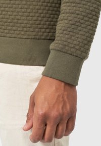 Gros plan sur la main d'une personne reposant le long du corps, portant un pull vert olive texturé avec des poignets côtelés et un pantalon clair.