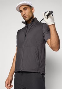 Man in witte pet, zwarte zip-up vest en golfhandschoen, met golfclub over zijn schouder, tegen een effen lichte achtergrond.