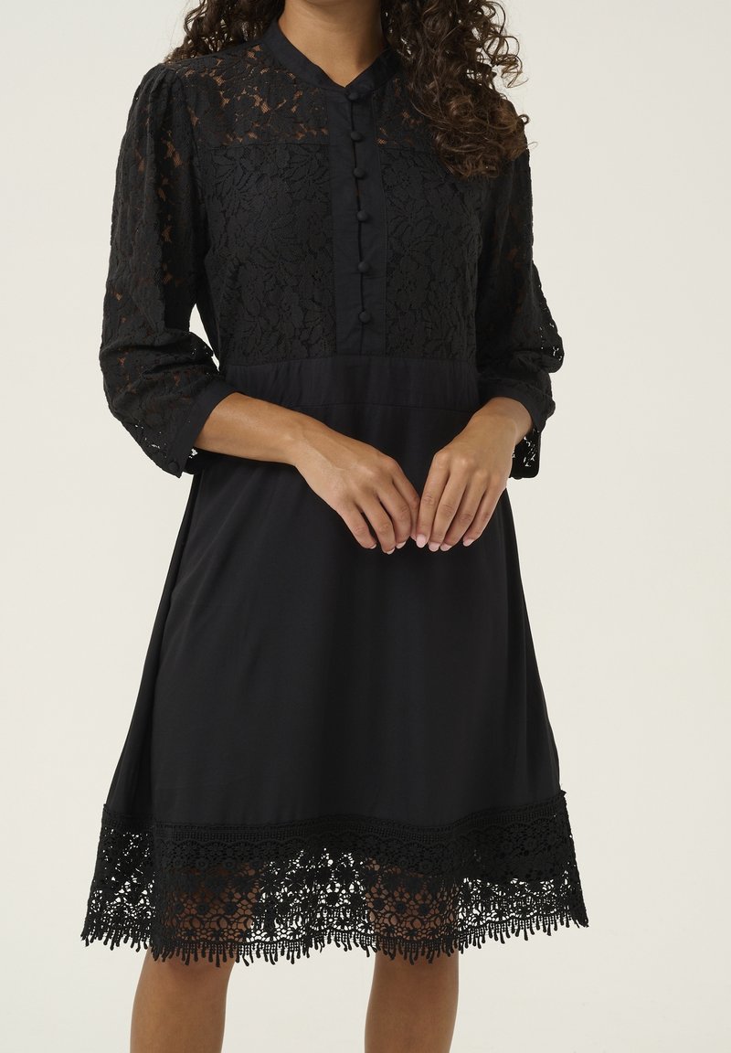 Femme portant une robe noire au genou avec des manches en dentelle, devant boutonné et ourlet en dentelle, debout les mains délicatement jointes.