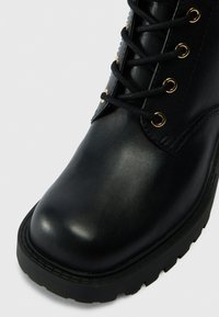 Botim preto de couro até ao tornozelo com biqueira arredondada, ilhós dourados e cordões pretos; apresenta uma sola grossa de borracha com padrões de tração.