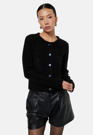 Mujer con cabello oscuro peinado hacia atrás, que lleva pendientes de aro plateados, un cárdigan negro con botones y pantalones cortos de cuero negros, de pie frente a un fondo blanco.