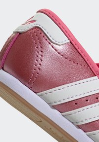 Talons de baskets brillants rose et blanc avec des rayures blanches, des coutures roses et une semelle marron clair sur un fond neutre.