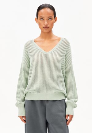 Pull en maille vert clair avec un col en V et des manches larges, présentant une texture côtelée et une coupe ample. Assorti à un pantalon gris.