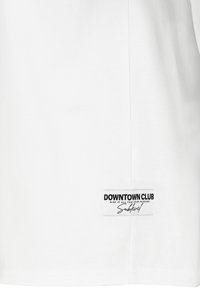 Weißes Baumwoll-T-Shirt mit kurzen Ärmeln, das an dem unteren Saum ein rechteckiges schwarzes Etikett mit "DOWNTOWN CLUB" und minimalem Text trägt.