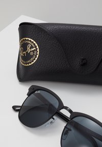 Occhiali da sole neri Ray-Ban con lenti scure, forma rettangolare e accenti laterali. Include una custodia in pelle nera con logo dorato in rilievo.