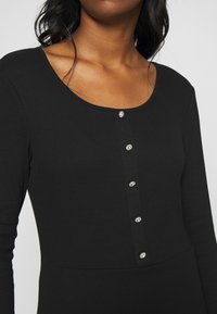 Svart ribbad långärmad topp med rundad halsringning och sex dekorativa knappar med klara detaljer nedtill. Slät textur.