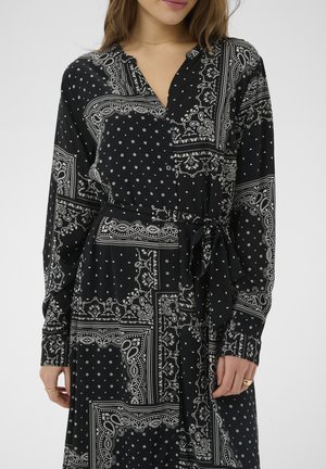 Kobieta ubrana w czarną sukienkę z długim rękawem z białymi wzorami paisley i kropkami, posiadająca dekolt w szpic i pasek wiązany w talii.