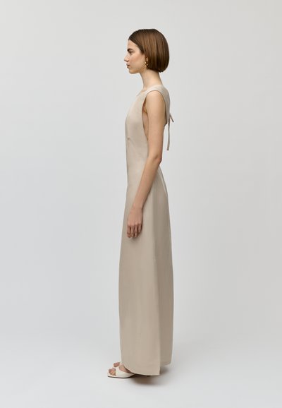 Beige, mouwloze maxi-jurk met een textuurafwerking en een strikdetail op de rug. Model draagt witte open teen hakken.