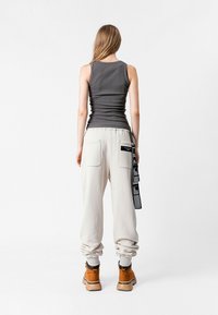 Camiseta sin mangas gris, pantalones de chándal beige con puños elásticos, dos bolsillos traseros y un detalle de etiqueta negra. Botas marrones completan el atuendo.