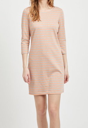 Kobieta ubrana w sukienkę sięgającą kolan, w poziome paski w kolorze brzoskwiniowym i beżowym, z rękawami do połowy przedramienia, na jednolitym jasnym tle.