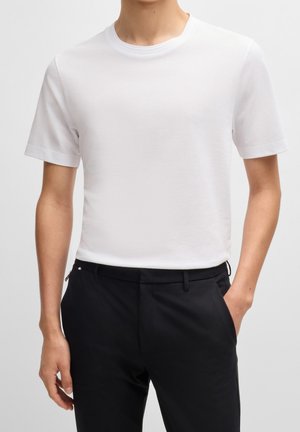 Man wearing short-sleeve white textured crew neck shirt tucked into black trousers with one hand in pocket, against plain background.