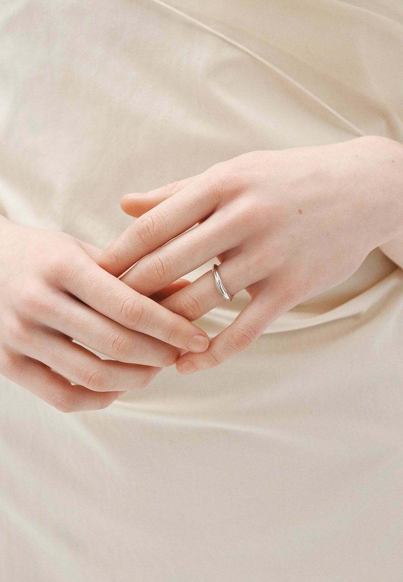 Anillo de plata con un diseño retorcido, llevado en un dedo, descansando entre manos de tono de piel claro sobre un fondo suave y neutro.