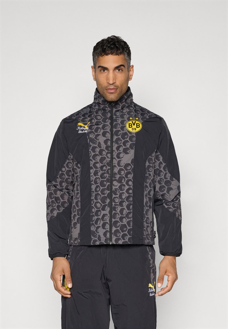 Schwarze Jacke mit strukturiertem grauem Muster, hohem Kragen, durchgehendem Reißverschluss und Logo auf der Brust. Kombiniert mit passenden schwarzen Hosen.