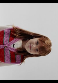 Jeune fille aux cheveux roux lisses portant un gilet en tricot rayé rouge et rose par-dessus une chemise blanche, face à l'avant sur un fond uni.