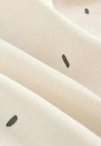 Hienokankainen puuvillakangas, jonka väri on kerma, ja siinä on eri kokoisia mustia pisteitä hajallaan pinnalla.