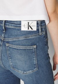 Džíny s vysokým pasem v modrém denimovém vzhledu s bledým texturováním, obsahující zadní kapsy a bílou etiketu s logem "Calvin Klein Jeans".