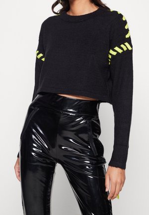 Kvinde iført sort cropped sweater med gule striber på ærmerne og skinnende sorte højtaljede bukser med lynlåslommer.