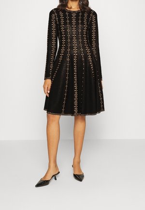 Kobieta ubraną w czarną sukienkę do kolan z długimi rękawami i geometrycznymi wycięciami, do tego czarne czółenka na niskim obcasie ze spiczastym noskiem.