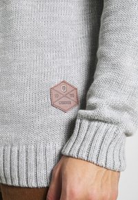 Grijze gebreide trui met geribbelde mouwen, voorzien van een bruine leren hexagonale patch met het label "INDICODE 1993."