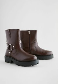 Botas de tobillo de cuero marrón con cremallera, detalle de hebilla y suela de goma negra gruesa. Presentan un acabado texturizado y costuras reforzadas.