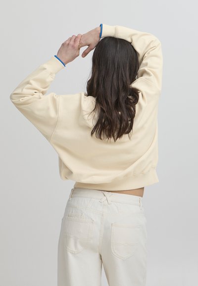 Sudadera color crema con hombros caídos y puños azules, combinada con pantalones blancos de cintura alta que tienen bolsillos traseros y detalles de costura sutil.