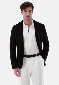 Blazer in velluto a coste marrone sopra una camicia bianca a righe verticali con bottoni scuri, abbinato a pantaloni bianchi e una cintura nera.
