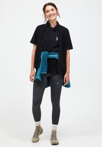 Schwarze Fleece-Weste mit Kragen über einem schwarzen T-Shirt, kombiniert mit dunklen Leggings und beigen Wanderschuhen, akzentuiert durch einen türkisfarbenen Pullover, der um die Taille gebunden ist.