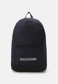 Sac à dos navy avec base noire, poche avant zippée et texte "HILFIGER" blanc. Dispose d'une poignée supérieure et de bretelles rayées.