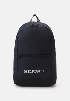 Zaino navy con base nera, tasca anteriore con zip e testo bianco "HILFIGER". Presenta una maniglia superiore e spallacci a righe.