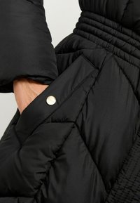 Melnā pūkainā jaka ar quilted dizainu, kuru papildina sānu kabata ar metāla sprādzi un ribota josta labākai piegustei.