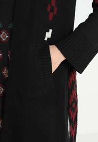 Cardigan nero lavorato a maglia con motivi geometrici rossi e bianchi, polsini a coste e tasche frontali. Il tessuto testurizzato aggiunge calore.