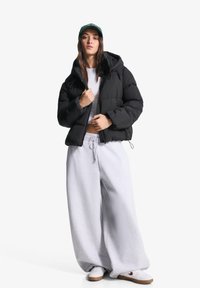 Giubbotto imbottito nero con cappuccio, top corto beige e pantaloni larghi grigi in felpa. Sneakers bianche con suole marroni. Cappellino verde con testo.