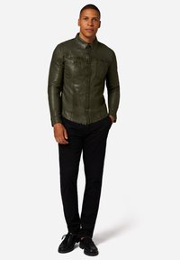 Olivgrön, figursydd skjorta med knäppning och spridd krage, två bröstfickor och långa ärmar, samt svarta byxor och svarta skor.