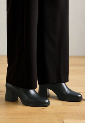 Crne kožne čizme s potpeticom i okruglim vrhom, uparene s opuštenim crnim hlačama, na pozadini drvenog poda.