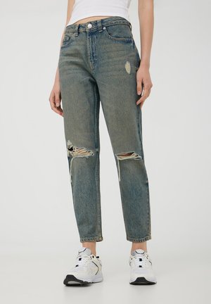 Jeans in denim azzurro chiaro con vestibilità rilassata, dettagli strappati sul ginocchio, cuciture a vista e design a cinque tasche. Abbinati a sneakers bianche.