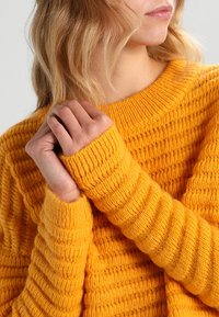 Maglione giallo brillante lavorato a maglia con texture a costine e un ampio scollo. I polsini sono aderenti, con strisce verticali che evidenziano calore e comfort.