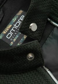 Grön texturerad tyg med ett vävt mönster, med silverfärgad hårdvara och en etikett som säger "ombre" med en blommig design. Dragkedjedetalj ingår.