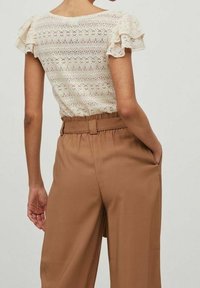 Blusa di pizzo color crema con maniche corte a volant, abbinata a pantaloni a vita alta e dalla vestibilità ampia in marrone chiaro, dotati di tasche laterali.