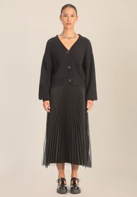 Czarny kardigan o teksturowanej fakturze z dekoltem w serek, zapinany na guziki, z oversize'owymi rękawami, w połączeniu z czarną spódnicą plisowaną i butami na koturnie.