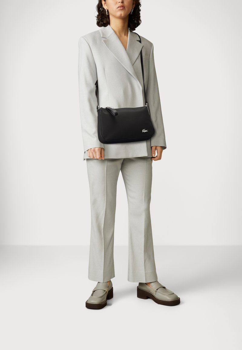 Bolso de hombro de cuero negro con una textura suave, forma redondeada, herrajes plateados y un logo sutil. Lleva puesto un traje gris claro y zapatos.