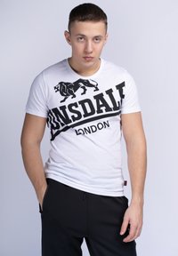 Baltas trumpomis rankovėmis marškinėliai su juodu liūto piešiniu ir ryškiu "LONSDALE LONDON" užrašu per krūtinę. Jaučiami su juodomis kelnėmis.