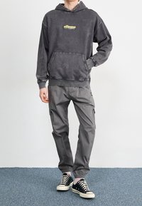 Sudadera con capucha gris descolorida con logo amarillo, combinada con pantalones cargo grises y zapatillas negras bajas. Tela texturizada y ajuste holgado.