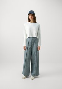 Hvit cropped langermet topp, lyseblå vide bukser med vertikale folder, og en mørk caps. Modell som har på seg hvite joggesko.