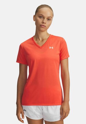 Vrouw die een feloranje V-hals Under Armour T-shirt en witte mesh shorts draagt, staand tegen een effen lichte achtergrond.