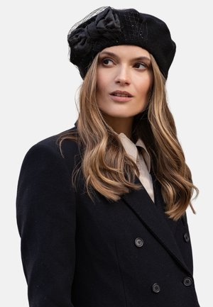 Berretto berretto nero con dettagli decorativi a fiocchi e rete, indossato con un cappotto doppio petto nero su una camicia bianca.