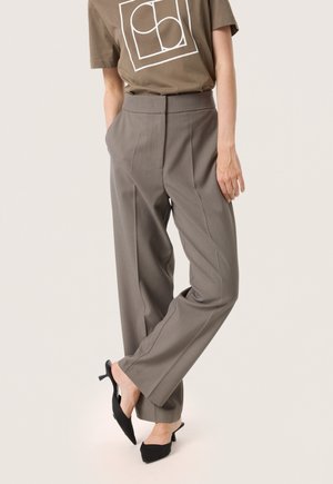 Persona que lleva pantalones taupe de cintura alta con pliegues frontales y bolsillos, combinados con una camiseta gráfica marrón y tacones slingback puntiagudos negros.