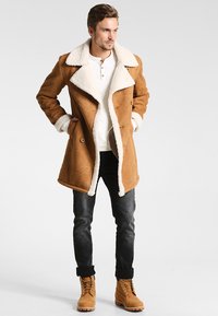 Hombre con abrigo de borrego marrón, camiseta henley blanca, jeans negros y botas de cordones beige, de pie con las manos en los bolsillos.