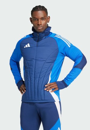 Veste de sport bleue avec un col haut, texture matelassée et rayures blanches sur les manches. Présente une fermeture éclair à l'avant et une coupe ajustée.