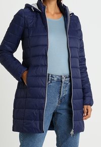 Abrigo acolchado azul marino con capucha ajustable, diseño acolchado, cremallera gris y bolsillos laterales. Lleva puesto encima una camiseta azul claro y pantalones vaqueros.